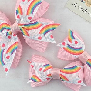May include: Three pink hair bows with a white background featuring a rainbow and heart design.  The bows are different sizes.