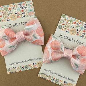 May include: Two peach-patterned hair bows with pink centers. The bows are displayed on white cards with the text "JL Craft & Design" and a website address.