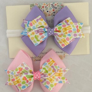 Pu&ograve; includere: Due fiocchi per capelli a tema pasquale con un elastico bianco. I fiocchi sono realizzati in nastro viola e rosa con sfondo bianco e un motivo di uova colorate, coniglietti e fiori. L'elastico &egrave; bianco e ha un motivo a pois bianchi.