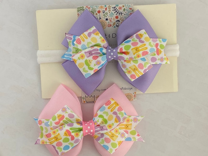 Pu&ograve; includere: Due fiocchi per capelli a tema pasquale con un elastico bianco. I fiocchi sono realizzati in nastro viola e rosa con sfondo bianco e un motivo di uova colorate, coniglietti e fiori. L'elastico &egrave; bianco e ha un motivo a pois bianchi.