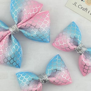 May include: Three mermaid-themed hair bows in different sizes. The bows are made of pink and blue fabric with a silver glitter overlay. The bows have a scale pattern and are tied with a silver glitter ribbon.
