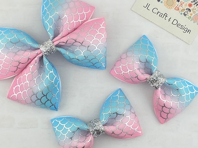 May include: Three mermaid-themed hair bows in different sizes. The bows are made of pink and blue fabric with a silver glitter overlay. The bows have a scale pattern and are tied with a silver glitter ribbon.