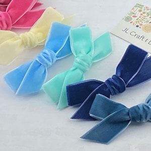 May include: A set of six velvet hair bows in various pastel colors, including light blue, yellow, pink, mint green, and two shades of blue. The bows are arranged in a row on a white surface. JL Craft & Design is printed on a card in the background.