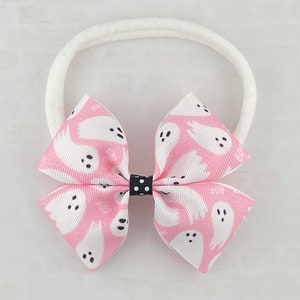 May include: A pink and white headband with a bow featuring a ghost pattern. The bow has black polka dots in the center.