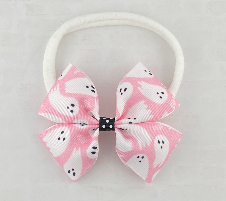 May include: A pink and white headband with a bow featuring a ghost pattern. The bow has black polka dots in the center.
