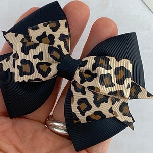 Peut inclure: Un n&oelig;ud pour cheveux fait &agrave; la main avec une base noire et un ruban &agrave; imprim&eacute; l&eacute;opard superpos&eacute;. Le n&oelig;ud est tenu dans une main, mettant en valeur les couleurs et les textures contrast&eacute;es. Un accessoire &eacute;l&eacute;gant.