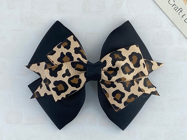 Peut inclure: Un n&oelig;ud de cheveux noir avec un imprim&eacute; l&eacute;opard. Le n&oelig;ud pr&eacute;sente un design superpos&eacute; avec une base noire unie et un centre imprim&eacute; l&eacute;opard beige et marron, adapt&eacute; &agrave; diverses occasions.