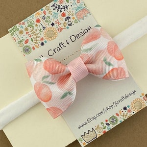 May include: A white headband with a pink bow featuring a peach print. The bow is tied with a pink ribbon. The headband is on a card with the text "Jill Craft & Design" and "www.Etsy.com/shop/jillcraftdesign".