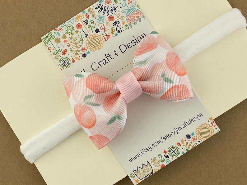 May include: A white headband with a pink bow featuring a peach print. The bow is tied with a pink ribbon. The headband is on a card with the text "Jill Craft & Design" and "www.Etsy.com/shop/jillcraftdesign".