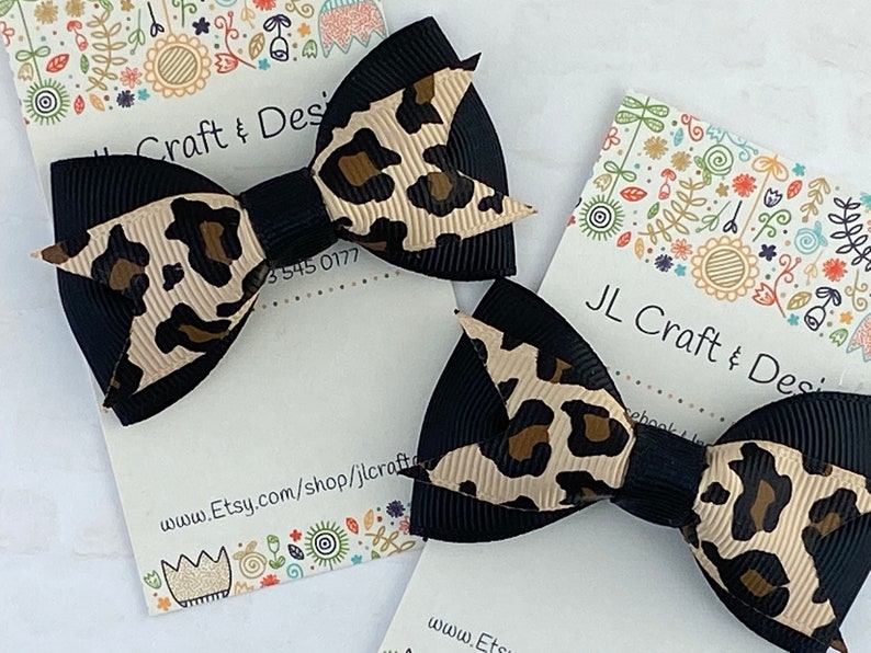 Peut inclure: Deux n&oelig;uds pour cheveux noirs et imprim&eacute; l&eacute;opard beige et marron. Les n&oelig;uds sont sur une carte blanche avec le texte "JL Craft & Design" et l'URL de la boutique Etsy. Ruban noir et imprim&eacute; l&eacute;opard beige et marron.