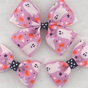 May include: Three purple and white hair bows with a ghost and candy corn pattern. The bows have black and white polka dot centers.