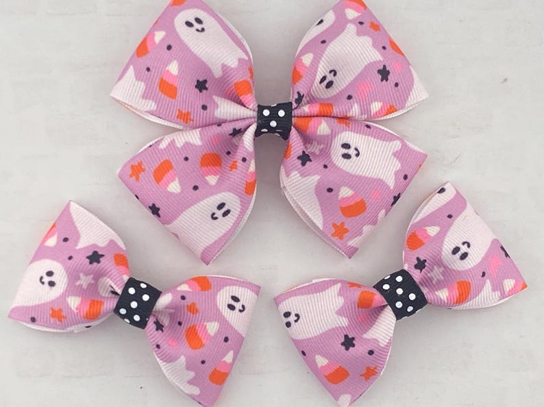 May include: Three purple and white hair bows with a ghost and candy corn pattern. The bows have black and white polka dot centers.