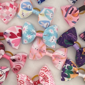 May include: A collection of colorful hair bows with various patterns, including hearts, bunnies, flowers, and Mardi Gras masks. The bows are made of fabric and have elastic bands.
