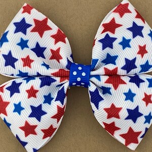 May include: A white bow with red and blue stars printed on it. The bow is tied with a blue ribbon with white polka dots.