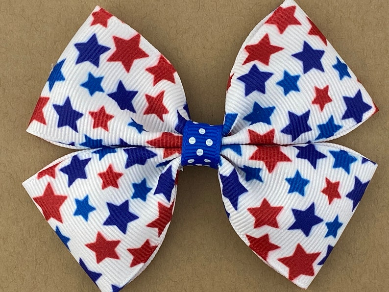 May include: A white bow with red and blue stars printed on it. The bow is tied with a blue ribbon with white polka dots.