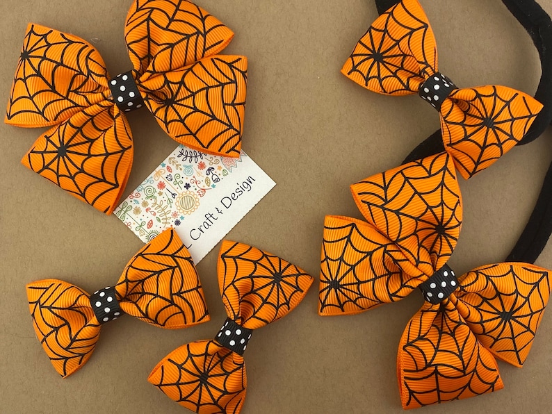 Puede incluir: Conjunto de cuatro lazos para el pelo de color naranja y negro con un patr&oacute;n de telara&ntilde;a. Los lazos est&aacute;n atados con una cinta de lunares negros y blancos. Los lazos est&aacute;n sobre un fondo marr&oacute;n.