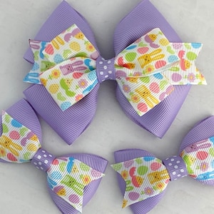 Pu&ograve; includere: Tre fiocchi per capelli a tema pasquale in viola e bianco con disegni di coniglietti e uova. Il fiocco pi&ugrave; grande &egrave; un fiocco a doppio strato con un retro viola. I due fiocchi pi&ugrave; piccoli sono a strato singolo.