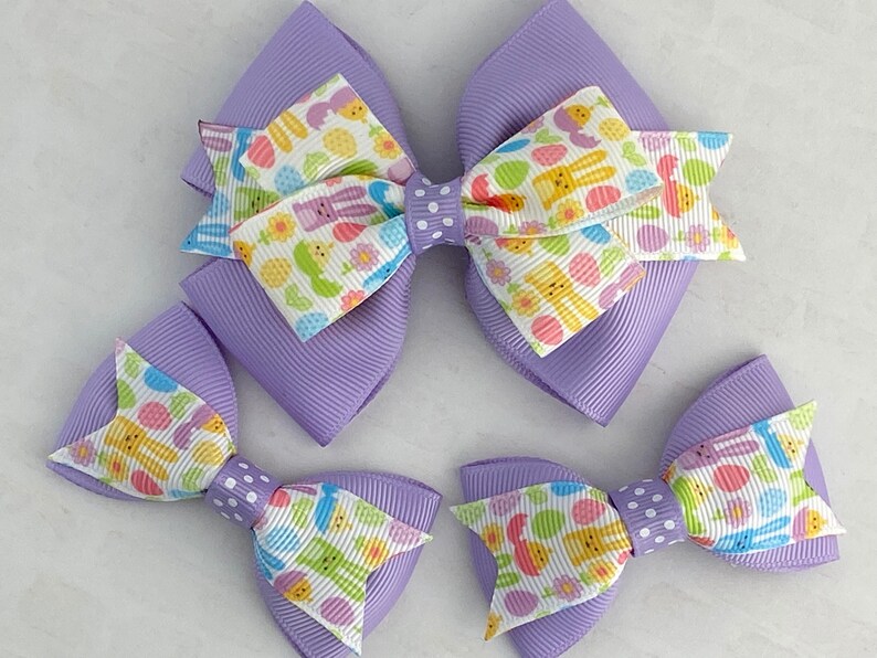 Pu&ograve; includere: Tre fiocchi per capelli a tema pasquale in viola e bianco con disegni di coniglietti e uova. Il fiocco pi&ugrave; grande &egrave; un fiocco a doppio strato con un retro viola. I due fiocchi pi&ugrave; piccoli sono a strato singolo.