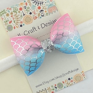 May include: A pink and blue ombre bow with a silver glitter center, attached to a white elastic headband. The bow has a mermaid scale pattern. The headband is on a white card with the text "Craft & Design" and a website address.