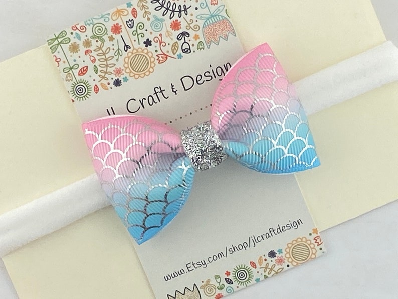May include: A pink and blue ombre bow with a silver glitter center, attached to a white elastic headband. The bow has a mermaid scale pattern. The headband is on a white card with the text "Craft & Design" and a website address.