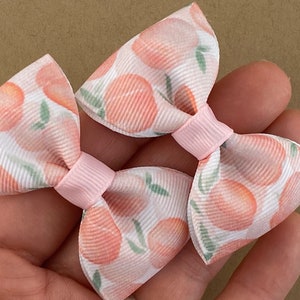 May include: Two peach-patterned hair bows with pink centers. The bows are made of white fabric with a peach print and have a pink center.