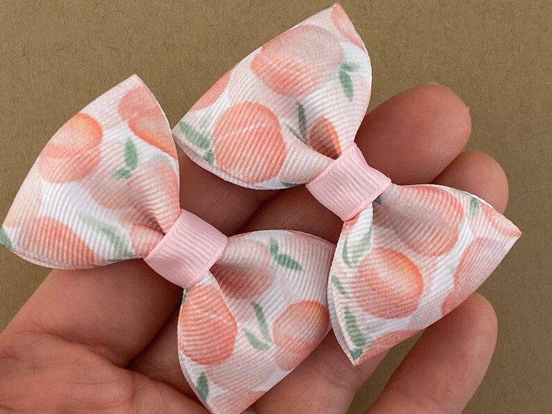 May include: Two peach-patterned hair bows with pink centers. The bows are made of white fabric with a peach print and have a pink center.