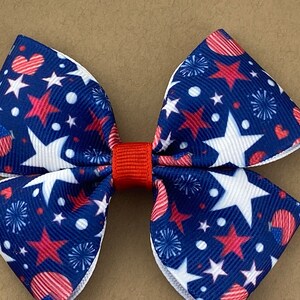 May include: A red, white, and blue bow with a stars and fireworks pattern. The bow is made of fabric and has a red center.