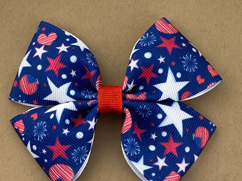 May include: A red, white, and blue bow with a stars and fireworks pattern. The bow is made of fabric and has a red center.