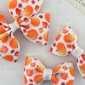 May include: Three white hair bows with orange and white pumpkin and leaf print. The bows are different sizes.