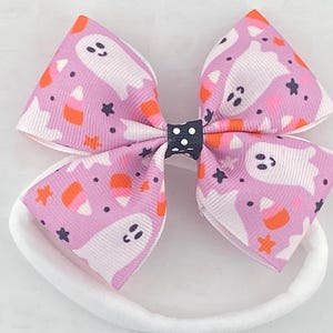 May include: A pink and white headband with a bow featuring a Halloween print of ghosts and candy corn. The bow is tied with a black and white polka dot ribbon.