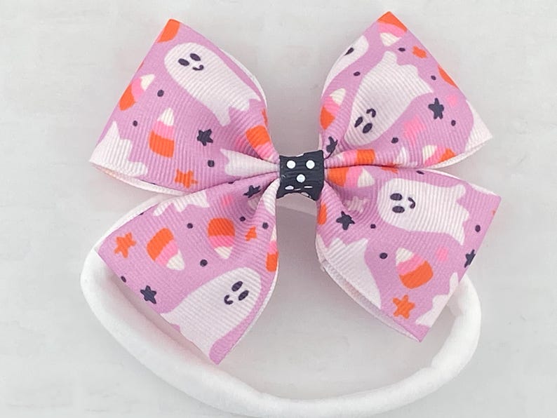 May include: A pink and white headband with a bow featuring a Halloween print of ghosts and candy corn. The bow is tied with a black and white polka dot ribbon.