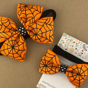 Puede incluir: Dos lazos para el pelo de color naranja y negro con un patr&oacute;n de telara&ntilde;a. Los lazos est&aacute;n unidos a gomas para el pelo negras. El lazo m&aacute;s peque&ntilde;o est&aacute; en una tarjeta con el texto "www.Etsy.com/shop/"