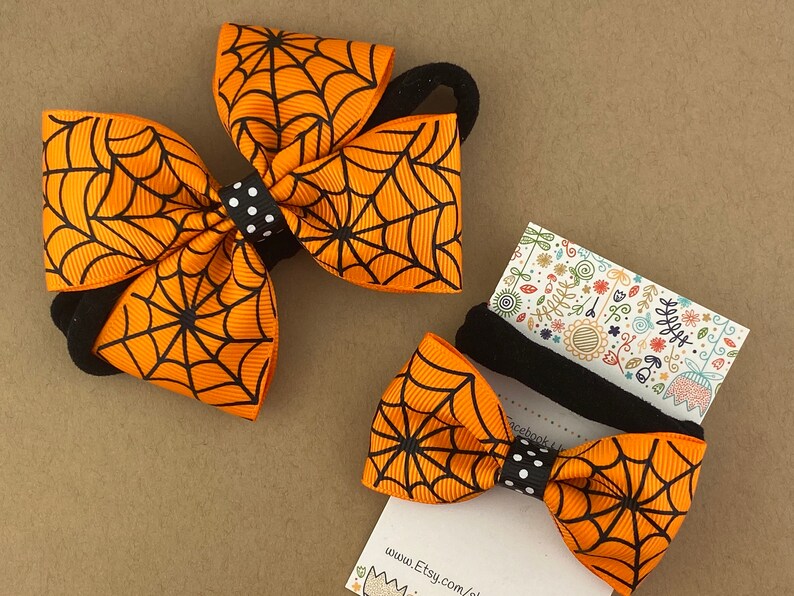 Puede incluir: Dos lazos para el pelo de color naranja y negro con un patr&oacute;n de telara&ntilde;a. Los lazos est&aacute;n unidos a gomas para el pelo negras. El lazo m&aacute;s peque&ntilde;o est&aacute; en una tarjeta con el texto "www.Etsy.com/shop/"
