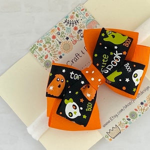 May include: Orange and black Halloween hair bow with a white polka dot pattern. The bow features a ghost and skull design with the text "cute spook" and "boo!"