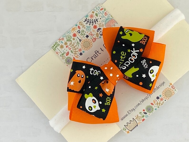 May include: Orange and black Halloween hair bow with a white polka dot pattern. The bow features a ghost and skull design with the text "cute spook" and "boo!"