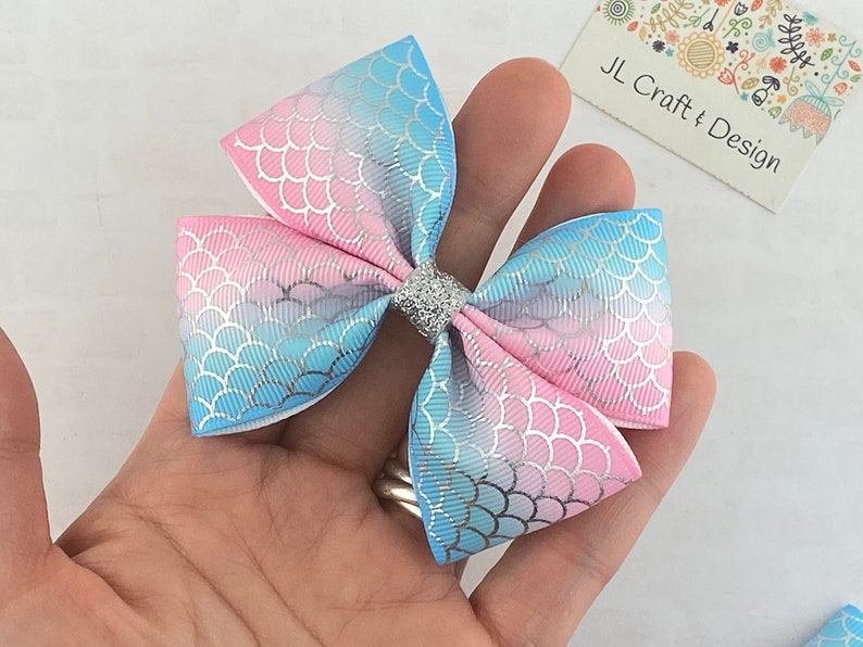 May include: A blue and pink ombre hair bow with a mermaid scale pattern and a silver glitter center.  JL Craft & Design.