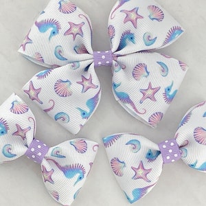 May include: Three white hair bows with a purple and blue seahorse, seashell, and starfish pattern. The bows have a purple polka dot center.