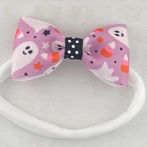 May include: A white headband with a purple and white bow featuring a ghost and candy corn pattern. The bow has a black and white polka dot center.