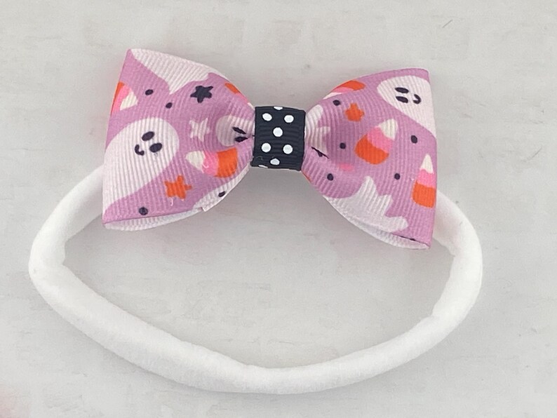May include: A white headband with a purple and white bow featuring a ghost and candy corn pattern. The bow has a black and white polka dot center.