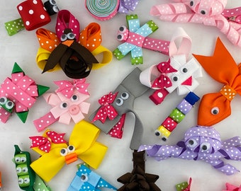 Può includere: Una collezione di fermagli per capelli colorati realizzati in nastro gros-grain, con varie forme di animali e personaggi dotati di occhi mobili. L'assortimento include disegni come un tacchino, un maiale, un polpo e un bruco a pois e tinta unita.