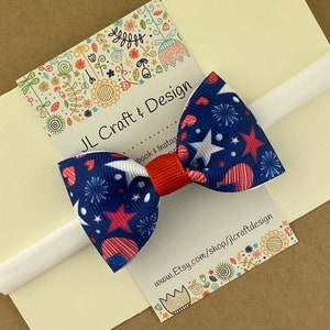 May include: A red, white, and blue bow with stars and hearts on a white headband. The bow is attached to a card that says "JL Craft & Design" and has a website address.