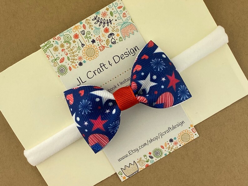 May include: A red, white, and blue bow with stars and hearts on a white headband. The bow is attached to a card that says "JL Craft & Design" and has a website address.