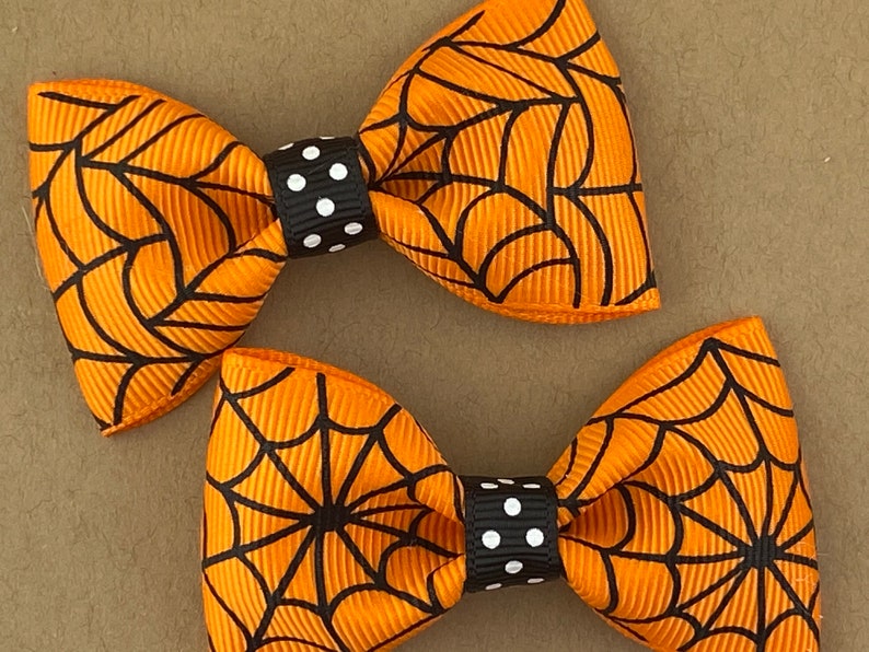 Puede incluir: Dos lazos para el pelo de color naranja y negro con estampado de telara&ntilde;a y centros de lunares negros y blancos.
