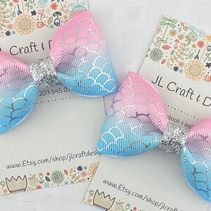 May include: Two pastel blue and pink hair bows with a silver glitter center. The bows have a mermaid scale pattern.