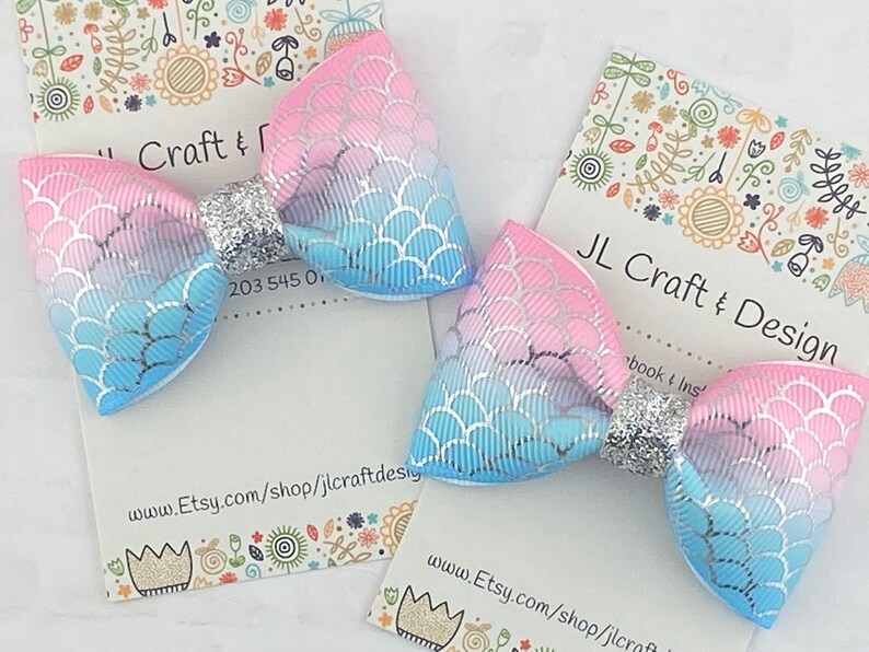 May include: Two pastel blue and pink hair bows with a silver glitter center. The bows have a mermaid scale pattern.
