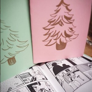 Puede incluir: Dos tarjetas de felicitaci&oacute;n, una verde menta y otra rosa, cada una con un dise&ntilde;o de &aacute;rbol de Navidad marr&oacute;n. Un c&oacute;mic abierto con ilustraciones en blanco y negro est&aacute; en primer plano.