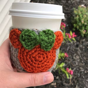 May include: A crocheted coffee cup cozy with a gray background, orange ears, and a green bow.