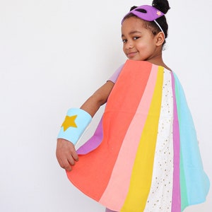 May include: A superhero costume set featuring a multi-coloured cape with vertical stripes in coral, orange, yellow, white with gold stars, purple, and light blue. The set includes a light blue wrist cuff with a yellow star and a purple eye mask.