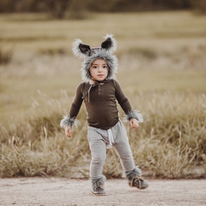 May include: A child in a wolf costume, featuring a brown long-sleeved top, grey trousers, and a furry hood with ears. The costume also includes furry cuffs and shoe covers. The child is standing on a dirt track in a field.