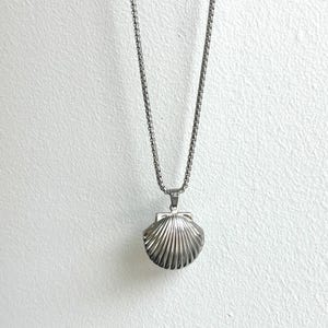May include: A silver-colored necklace with a seashell pendant. The pendant is a detailed, ridged seashell design. The necklace hangs against a white textured background. The text "SHOP MORNING BREAD" is at the bottom of the image.